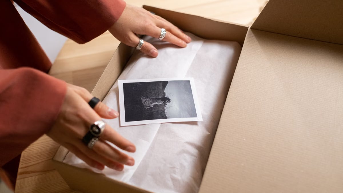 Hands opening a cardboard box revealing a photograph wrapped in tissue paper.