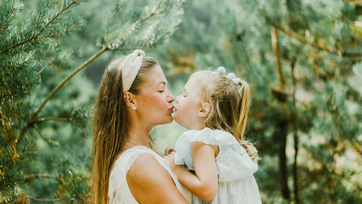A mother and daughter share a tender kiss surrounded by lush greenery, symbolizing love and bonding.